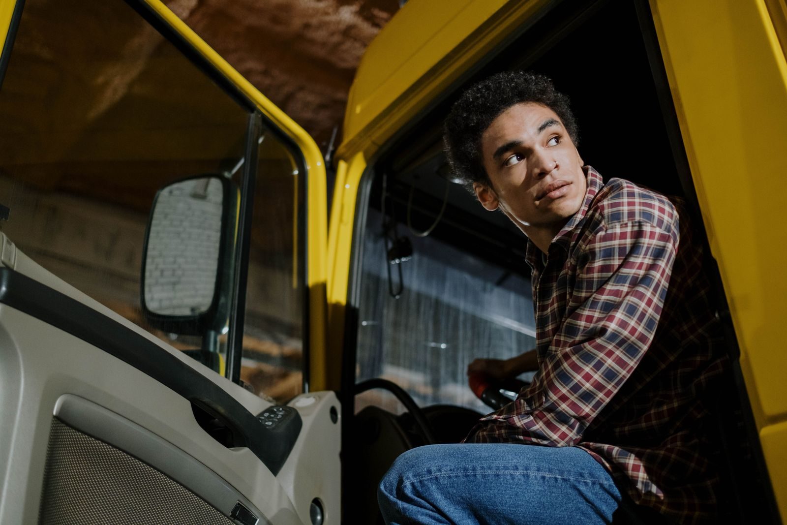 A driver in a plaid shirt sitting at the open door of his yellow semi-truck